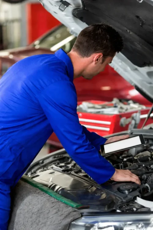 Ein Automechaniker in blauer Arbeitskleidung beugt sich über einen offenen Motorraum und arbeitet mit einem Tablet. Die Person ist von der Kamera abgewandt, Werkzeuge und eine rote Werkbank sind im Hintergrund sichtbar.